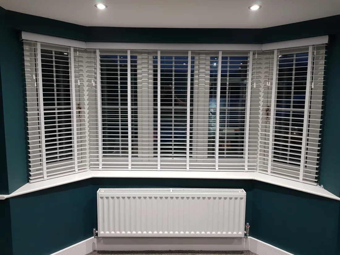 White faux wood blinds fitted in a bay window with deep teal walls